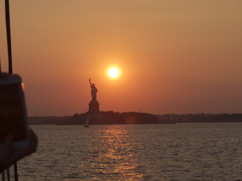 The sun going down over New York Harbor next to the Statue of Liberty.